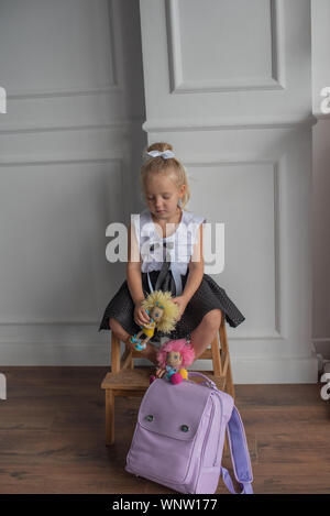 Close-up portrait of a little girl against a white wall.Girl in a school uniform with The girl holds dolls in her hands. A school backpack is on the f Stock Photo