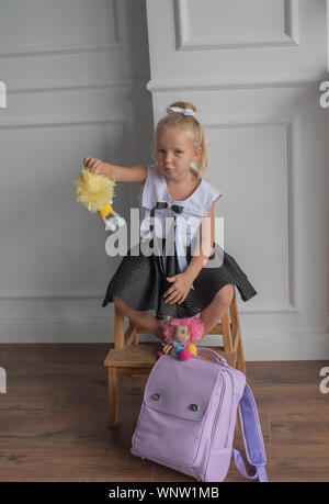 Close-up portrait of a little girl against a white wall.Girl in a school uniform with The girl holds dolls in her hands. A school backpack is on the f Stock Photo