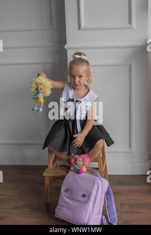 Close-up portrait of a little girl against a white wall.Girl in a school uniform with The girl holds dolls in her hands. A school backpack is on the f Stock Photo