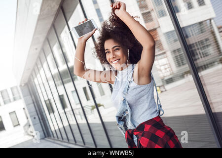 Young woman in the city street near window with smartphone up wearing earphones listening to music dancing cheerful Stock Photo