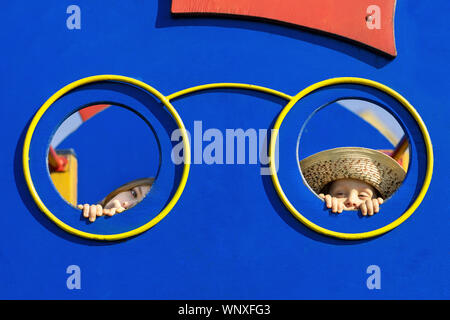 Boy and girl looking through hole in the wall of playhouse. Two kids hiding behind the playhouse wall and peeking or peeping. Stock Photo