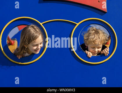 Two kids peeking through the hole on the playhouse. Boy and girl playing in park. Happy children concept. Stock Photo