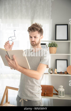 Handsome young caucasian Man reading a book in his living room ,feeling serious and surprised Stock Photo