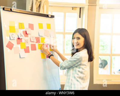 Happy young asian businesswoman with colored sticky notes ,posted notes on white board with erasable marker in office.Start up home business informati Stock Photo