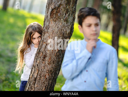 Boy and girl playing hide and seek in the park. Girl watching on boyfriend. Girl hiding behind the tree and lurk on boy. Stock Photo