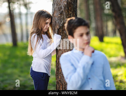 Boy and girl playing hide and seek in the park. Girl watching on boyfriend. Girl hiding behind the tree and lurk on boy. Stock Photo