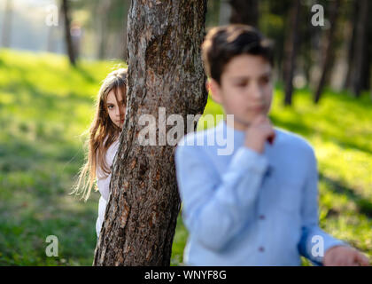 Boy and girl playing hide and seek in the park. Girl watching on boyfriend. Girl hiding behind the tree and lurk on boy. Stock Photo