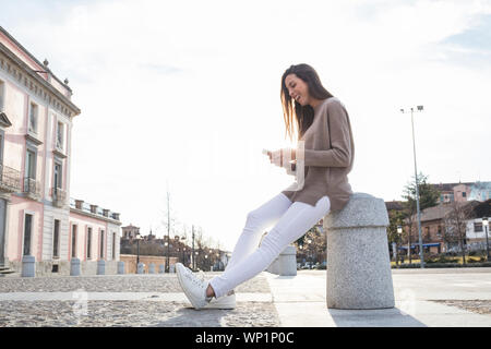 Smiling woman sitting on a bollard looking at the mobile Stock Photo