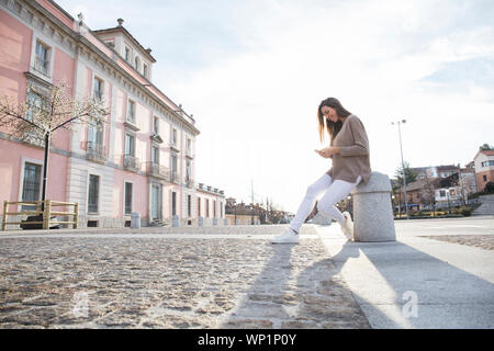 Smiling woman sitting on a bollard looking at the mobile in a street Stock Photo