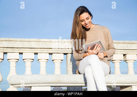 Smiling woman using her tablet while sitting on a stone bench Stock Photo