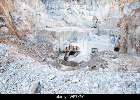 Excavator or digger inside an open cast mining pit for white Carrara marble showing large blocks of stone cut from the mountainside in Tuscany, Italy Stock Photo