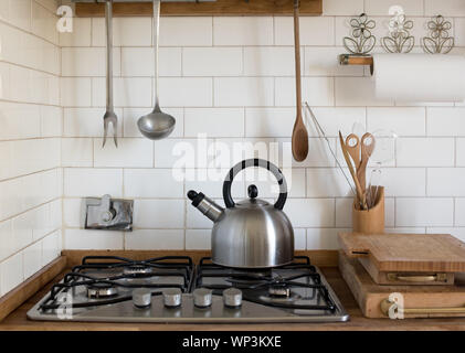 Stainless steel kettle standing on an unlit gas burner on the hob in a kitchen with tiled white splash back and hanging utensils Stock Photo