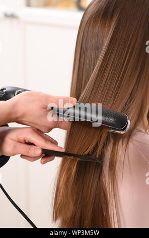 Close up on unidentifiable persons hands using black hair straightener and comb on womans brown hair Stock Photo