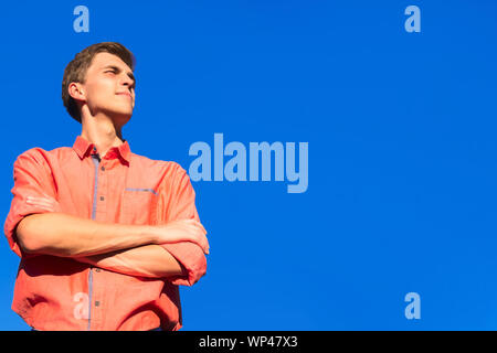 Serious cool young man in orange shirt standing with arms crossed against blue sky background Stock Photo