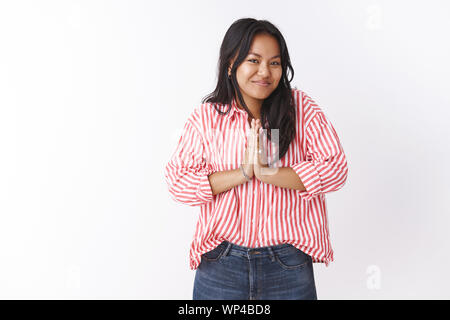 Namaste enjoy your stay. Portrait of cute pleasant and happy young nice vietnamese girl in striped blouse pressing palms together in asian greeting ge Stock Photo