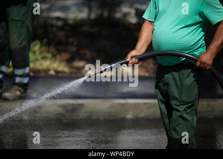 Man with big belly uses a hose to sprinkle water on the pavement while his colleagues sweep the street Stock Photo