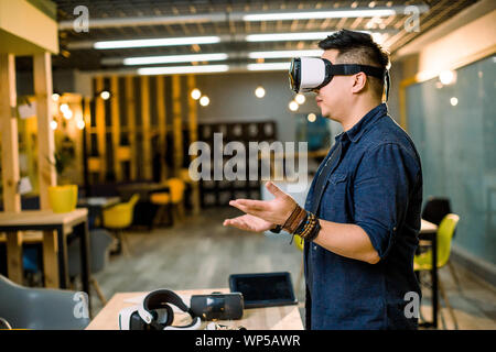 Future is now. Handsome young Chinese man in VR headset gesturing and smiling while standing in creative office Stock Photo