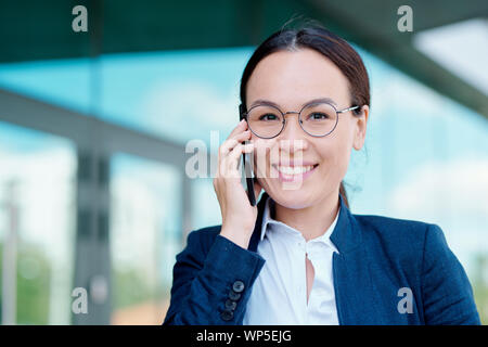 Elegant young female employee with toothy smile holding smartphone by ear Stock Photo