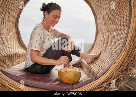 Young Woman Taking A Break And Drinking A Fresh Coconut Inside A Wooden Swing In Front Of The Sea, Bali, Indonesia Stock Photo