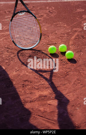 Still life of tennis balls and the shadow of a rman holding a tennis racquet on a red clay tennis court. Stock Photo