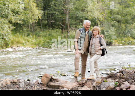 Affectionate mature spouses with backpacks standing on stones on river bank Stock Photo
