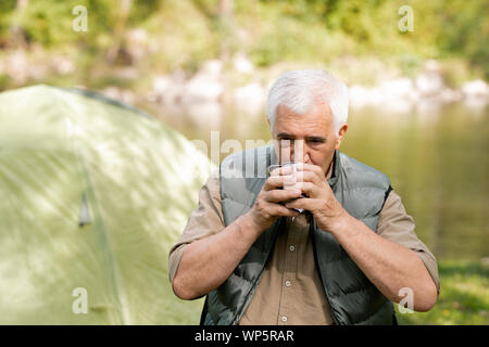 Senior hiker with grey hair drinking hot tea from tourist mug while having rest Stock Photo