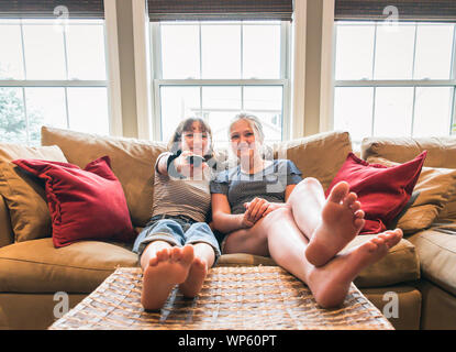 Two teenage girls sitting on a couch with feet up watching television. Stock Photo