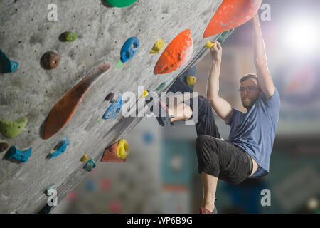 young active sport man with beard and glasses climbing on gym wall during bouldering workout Stock Photo