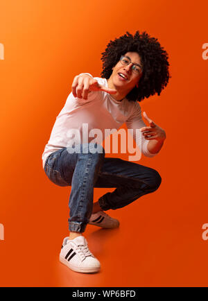 Cocky millennial guy pointing at camera, squatting on orange background Stock Photo