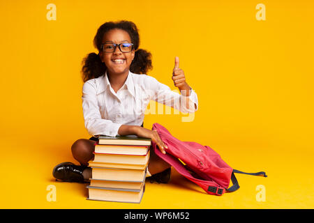 Afro Schoolgirl Gesturing Thumbs-Up Sitting At Book Stack, Studio Shot Stock Photo