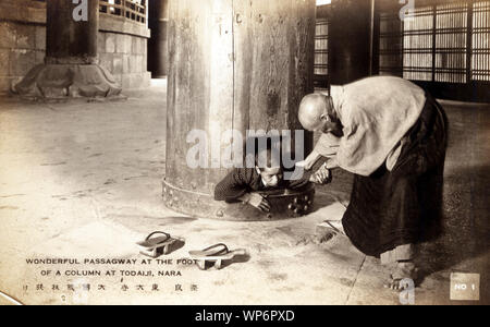 [ 1920s Japan - Interior of Todaiji in Nara ] —   A child crawls through a hole in a huge wooden pillar inside the buddhist temple Todaiji (Great Eastern Temple) in Nara, Japan.  It is believed that if you can crawl through, you will have a greater chance of attaining enlightenment.  The Great Buddha Hall of Todaiji is claimed to be the largest wooden building in the world. It houses a huge bronze statue of the Buddha, known as Daibutsu.  20th century vintage postcard. Stock Photo