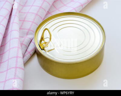 Yellow round tin can near checkered kitchen towel on a white table. Opening a food can with pull tab. Close-up. Stock Photo