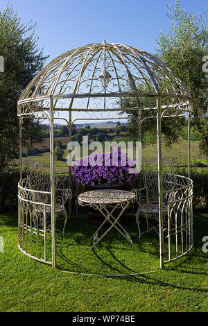 White wrought iron gazebo with table, chairs and purple flowers in a resort garden overlooking rolling hills of Tuscany, Italy Stock Photo