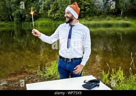An office worker holds a burning sparkler in his hand during a corporate party. White collar in a funny hat of a gnome, or Santa Claus, during the cel Stock Photo
