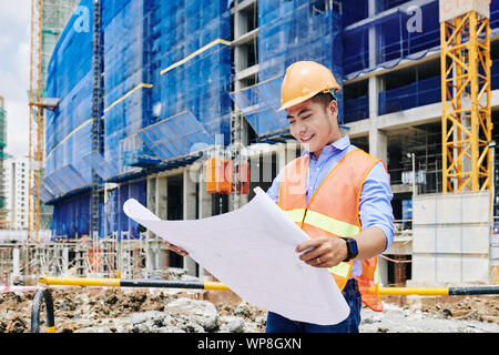 Smiling Asian construction engineer in orange vest and hardhat examining building blueprint Stock Photo
