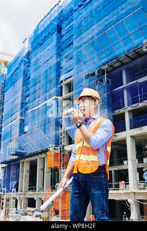 Young Vietnamese head engineer talking on walkie-talkie and giving orders to workers at construction site Stock Photo