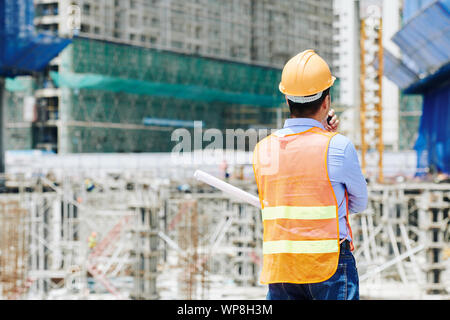 Rear view of professional engineer in orange vest using walkie-talkie to explain employees what to do Stock Photo