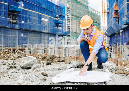 Professional Vietnamese engineer examining construction plan and talking on walkie-talkie with workers Stock Photo