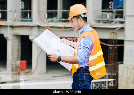 Smiling Vietnamese foreman examining blueprint of new building in his hands when working at construction site Stock Photo