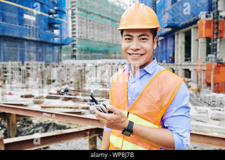 Portrait of cheerful Vietnamese construction engineer standing with portable radio communicator and smiling at camera Stock Photo