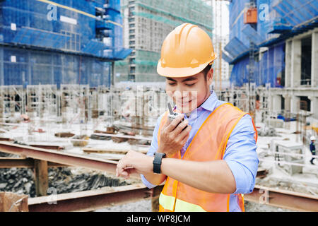 Smiling Asian contractor checking time on his wristwatch when talking to coworkers via walkie-talkie Stock Photo