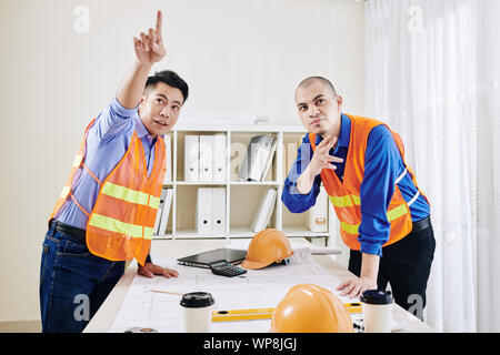 Multi-ethnic team of construction engineers leaning over table with building blueprint and pointing away at something interesting Stock Photo