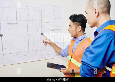 Serious multi-ethnic team of engineers discussing building blueprint hanging on the wall in architecture bureau Stock Photo