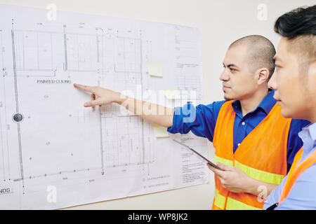 Serious engineer with digital tablet in hands pointing at building blueprint hanging on wall and discussing work with colleague Stock Photo