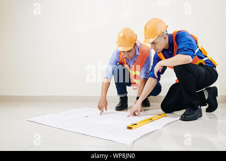 Engineers in hard hats and orange vests discussing floor layout in big blueprint on the floor Stock Photo