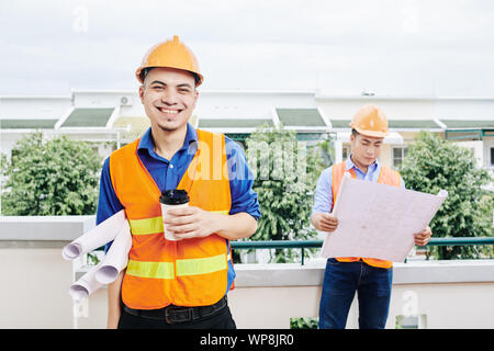 Smiling civil engineer standing outdoors with many blueprints scrolls and drinking cup of take away coffee, his coworker with building plan in backgro Stock Photo