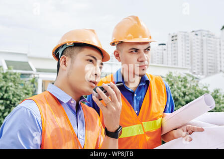 Vietnamese contractor talking on walkie-talkie with workers, his colleague standing nearby with blueprint in hands Stock Photo
