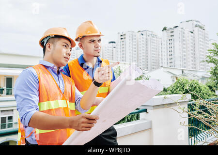 Serious construction workers standing outdoors with building blueprint and discussing sequence of work that needs to be done today Stock Photo