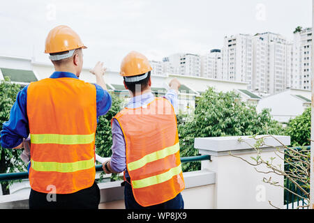 Rear view of civil engineers in bright orange vests pointing at building in new district they were designing Stock Photo