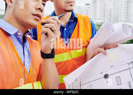 Cropped image of serious construction workers with building blueprint in hands talking on walkie-talkie with colleagues and controlling work Stock Photo